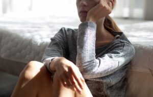 Image of a woman in a grey shirt sitting and staring off. Representing someone ho could benefit of meeting with a trauma therapist. Through trauma therapy and PTSD treatment you can move past this feeling and start feeling better.